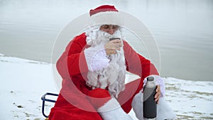 Santa Claus sits with a bag of presents on the snowy shore of a lake and drinks mulled wine. Drunk Santa Claus