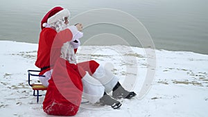 Santa Claus sits with a bag of presents on the snowy shore of a lake and drinks mulled wine. Drunk Santa Claus