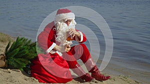 Santa Claus sits on the sandy shore of the lake sniffing the smell of alcohol and bottles. Drunk Santa Claus