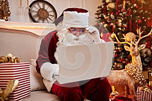 Santa Claus sitting at his home and reading email on laptop with ÃÂhristmas requesting or wish list near the fireplace and tree