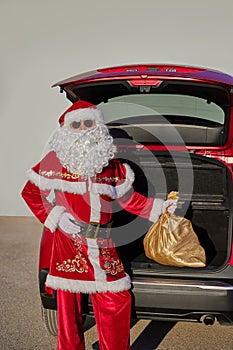 Santa Claus stands in front of an open car trunk and holds a bag of gifts in his hand
