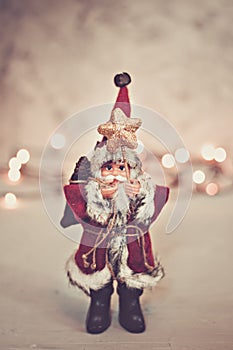 Santa Claus toy brings Christmas tree at blue snowy night bokeh background and blurred lights foreground.