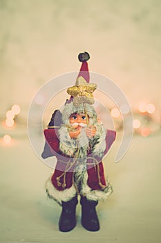 Santa Claus toy brings Christmas tree at blue snowy night bokeh background and blurred lights foreground.