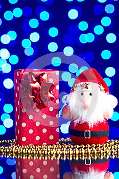 Santa Claus toy, gift in package and golden beads on a glass table with a beautiful blue bokeh