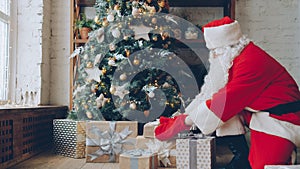 Santa Claus in traditional outfit putting gifts under Christmas tree on holiday at home