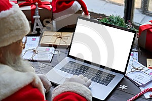 Santa Claus using laptop computer mock up white screen sitting at table.