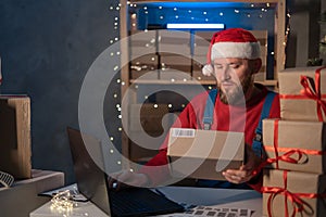 Santa Claus wearing hat holding gift box using laptop computer sitting at home office table late on Christmas eve