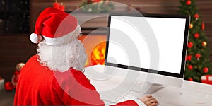 Santa Claus work on computer beside fireplace and Christmas tree