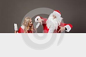Santa Claus and young beautiful mrs. Claus stand behind a white canvas and gesticulate on the gray background.