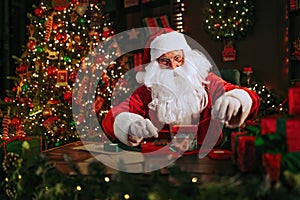 Santa Clause carefully is packing gifts while sitting at the table near Christmas tree. New Year concept