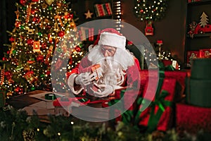 Santa Clause is packing gifts while sitting at the table near Christmas tree. New Year concept