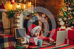 Santa helper baby boy with laptop. Bright New Years interior. Xmas tree. Indoor. Child at Christmas tree and fireplace