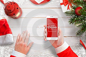 Santa with mobile phone on work desk. Christmas tree with decorations, gifts and lantern on white wooden desk