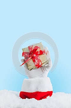 Santa's hand sticking out of a snowdrift. Santa Claus gloved hands holding gift box against light blue bokeh