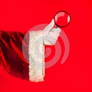 Santa`s hand in a traditional red suit and white glove holds a magnifying glass, a symbol of research, search, observation,