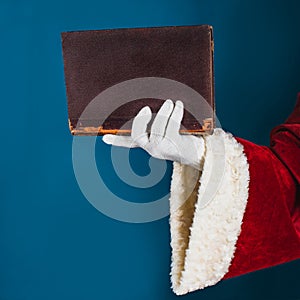 Santa\'s hand in a white glove holds a old book on a turquoise background with copy space. Concept for knowledge, education,