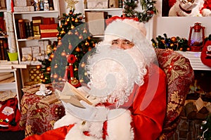 Santa sitting at the Christmas tree, near fireplace and reading a book