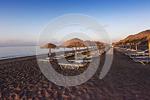 Santorini black beach Perissa, Perivolos illuminated by the morning rays of the sun. Beach chairs with umbrellas in a row behind