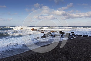 Santorini, the coast at Vlichada, the stormy sea crashes into the rocks at the coast. High waves, white foam and black sand on the