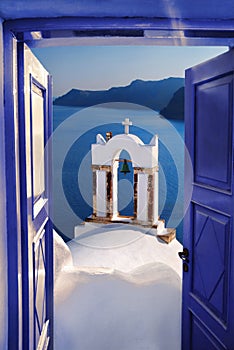 Santorini view with bell against open blue door in Oia village, Greece