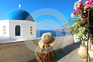 Santorini woman walking in beautiful street of Oia village with flowers and typical blue dome of the church, Santorini, Cyclades
