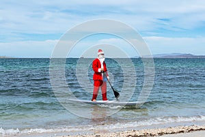 Santy Claus approaches the shore of the beach in his paddle boar. - Imagen