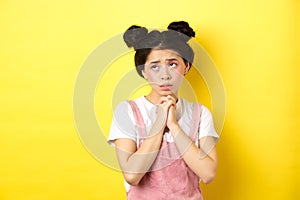 Sas asian girl holding hands in pray, pleading god, looking up and begging, standing on yellow background