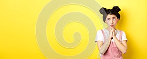 Sas asian girl holding hands in pray, pleading god, looking up and begging, standing on yellow background