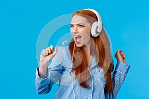 Sassy good-looking redhead female in pyjama having fun, listening music in big white headphones, singing into toothbrush