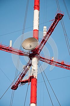 Satellite dishes and antennas on the red-white telecommunication tower against blue sky