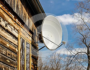 Satellite white dish on the wall under the roof of a wooden house against a blue spring sky and tree branches without leaves,