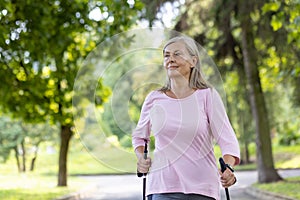 Satisfied and active senior gray-haired woman is doing sports and Nordic walking in the park, holding trekking poles in