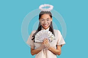 Satisfied angelic woman with nimb over head holding fan of dollars banknotes.