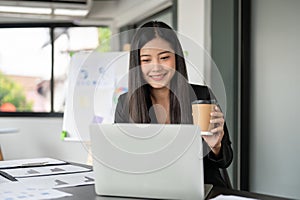 A satisfied Asian businesswoman is having a morning coffee while working on her laptop