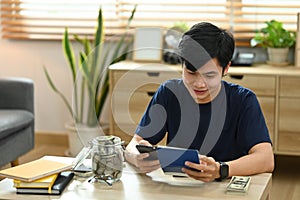 Satisfied asian man using smart phone for making online payments, managing expenses finances in living room