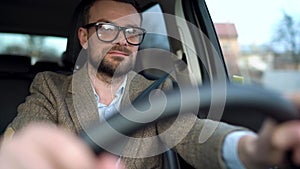 Satisfied bearded man in glasses driving a car down the street in sunny weather