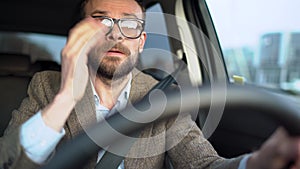Satisfied bearded man in glasses driving a car down the street in sunny weather