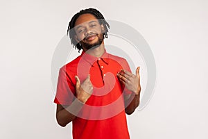 Satisfied bearded man wearing red casual style T-shirt, pointing finger at himself feeling proud and ambitious, boasting