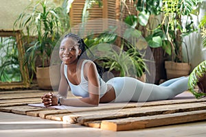 Satisfied black sported woman lying on fitness mat take break after yoga practice in greenery room.