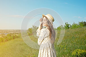 Satisfied brunette in a long summer dress wearing a straw hat is standing in the tall grass with her arms raised facing the