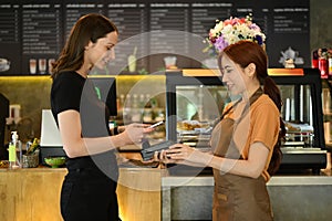 Satisfied caucasian female customer making contactless payment with smiling waitress in modern coffee shop