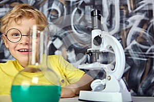Satisfied child in round glasses and T-shirt hides behind laboratory objects