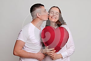 Satisfied delighted Caucasian man and woman holding heart shaped air balloon isolated over gray background handsome guy kissing