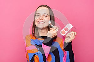 Satisfied delighted woman wearing sweater making makeup using cosmetic brush posing isolated on pink background, looking smiling