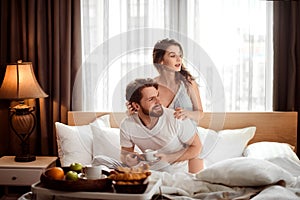 Satisfied family couple have breakfast in bed, enjoy delicious croissants, coffee and fruit, look positively away, sits