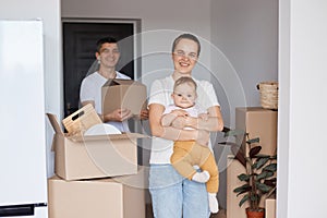 Satisfied family, mother, father and infant daughter posing in a new apartment after moving, expressing happiness surrounded with