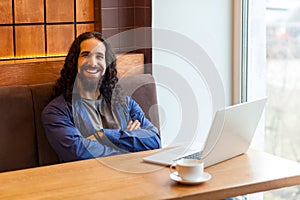 Satisfied handsome bearded young man freelancer in casual style sitting in cafe with laptop, crossing arms and looking at camera