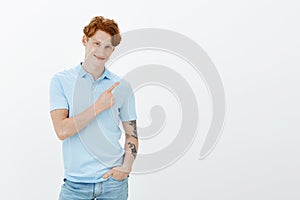 Satisfied handsome young male with red hair and freckles with tattoed arm, holding hand in pocket and pointing at upper