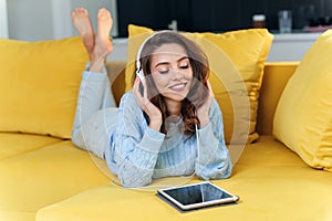 Satisfied happy girl using tablet while lying on comfortable yellow sofa and enjoying pleasant tunes with headphones.