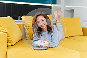 Satisfied happy girl using tablet while lying on comfortable yellow sofa and enjoying pleasant tunes with headphones.